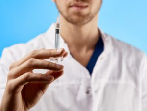 Doctor in white medical coat shows syringe to camera close-up isolated on blue background. Coronavirus vaccine, beauty injection concept